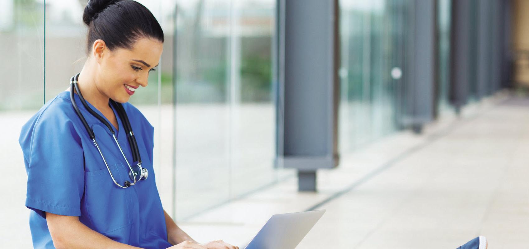 Nursing student in blue scrubs sitting against a window wall on her computer.